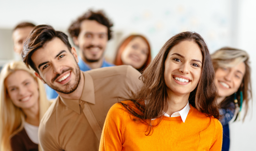 A group of cheerful, diverse young professionals standing in a diagonal line, leaning playfully toward the camera and smiling. The setting appears to be a bright, modern office space, suggesting a positive and collaborative team culture.