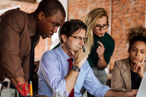 Four diverse professionals—two women and two men—gather around a laptop in a modern office with exposed brick walls. All appear focused and engaged, suggesting a collaborative work session or team discussion.