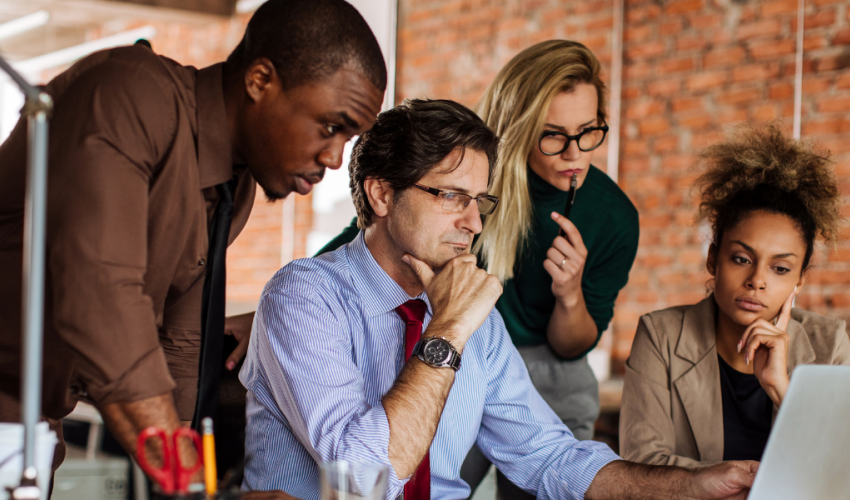 Four diverse professionals—two women and two men—gather around a laptop in a modern office with exposed brick walls. All appear focused and engaged, suggesting a collaborative work session or team discussion.